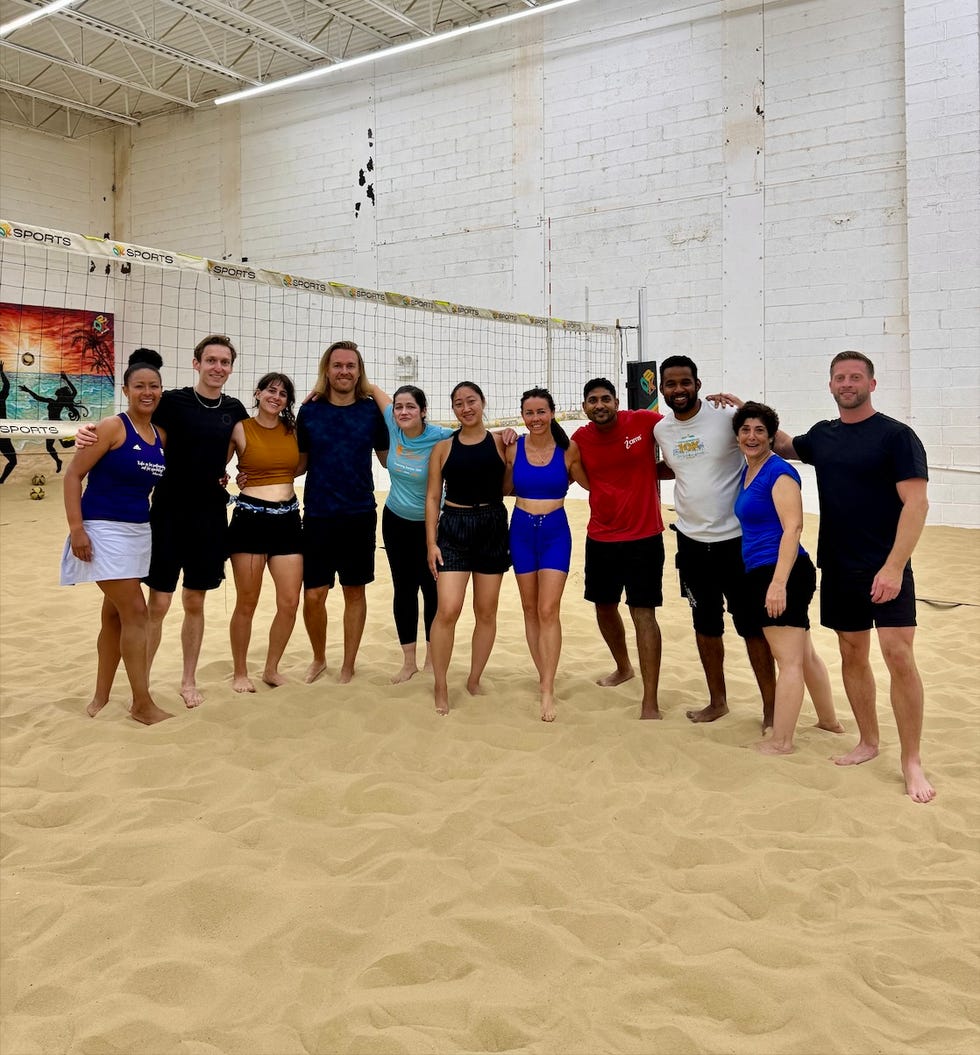 Group of people posing together on a sand volleyball court in an indoor facility.
