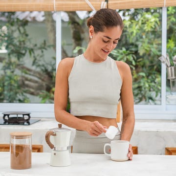 a woman adds a scoop of powder to her morning coffee in a bright, modern kitchen.