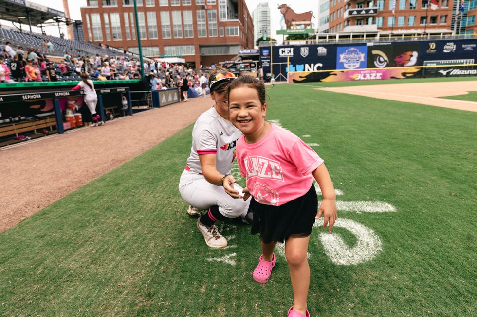 A smiling girl in a pink shirt and skirt stands on a baseball field, while an adult player kneels beside her. A smiling girl in a pink shirt and skirt stands on a baseball field, while an adult player kneels beside her.