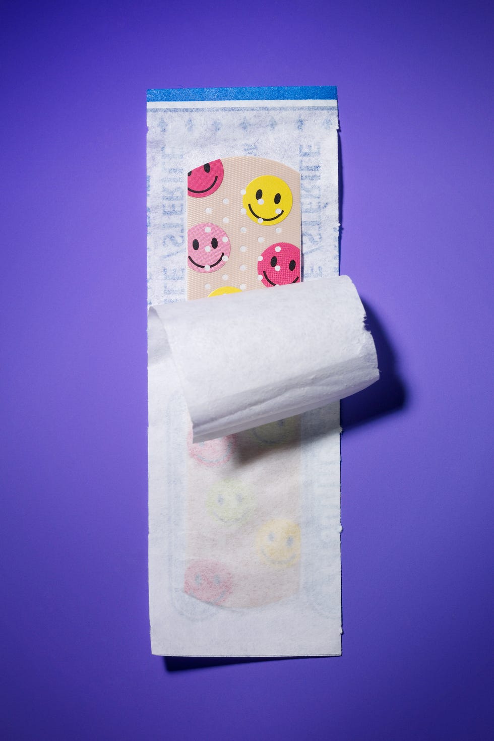 Colorful band-aid featuring smiley face designs on a purple background, partially unwrapped from its packaging. Colorful band-aid featuring smiley face designs on a purple background, partially unwrapped from its packaging.