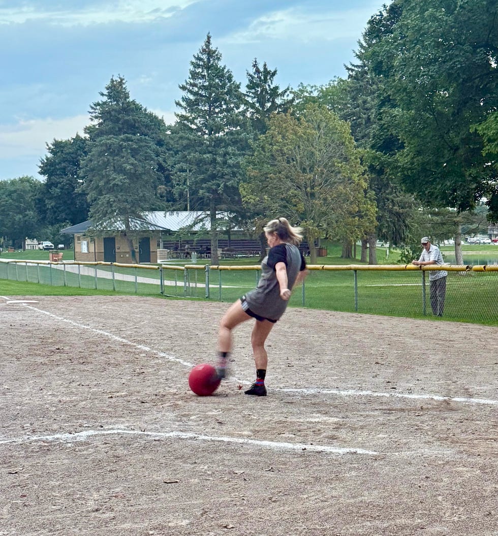 A girl kicks a red kickball on a gravel field surrounded by trees and park facilities.