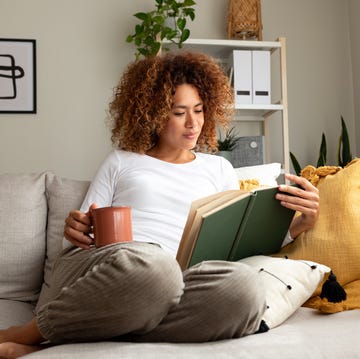 young multiracial woman reading a book at home, drinking tea sitting on the sofa in cozy living room. lifestyle concept.