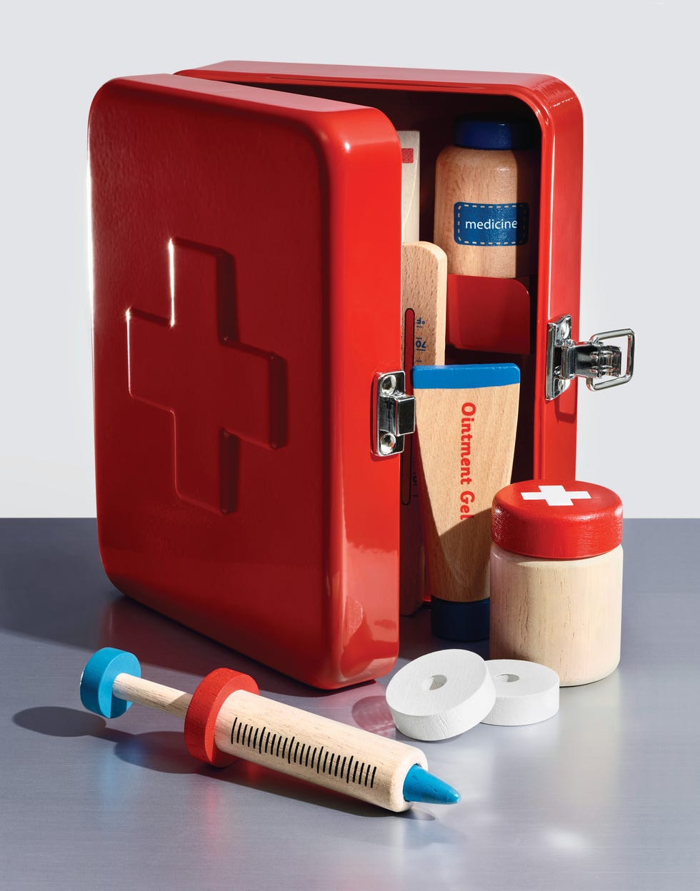 Toy red first aid kit with wooden medical supplies including tubes, a bottle, and a syringe on a silver surface. Toy red first aid kit with wooden medical supplies including tubes, a bottle, and a syringe on a silver surface.