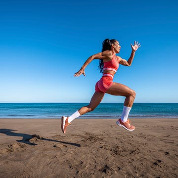 full length of muscular female runner in sportswear and sneakers jumping on sandy beach near waving sea against cloudless blue sky