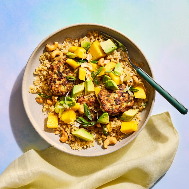 Bowl of quinoa topped with grilled patties, mango, avocado, green onions, and cashews, served with a fork and napkin.