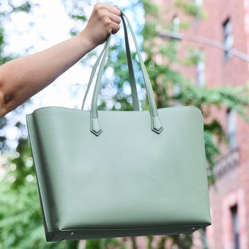 A hand holds a light green tote bag against an urban backdrop with trees and buildings.
