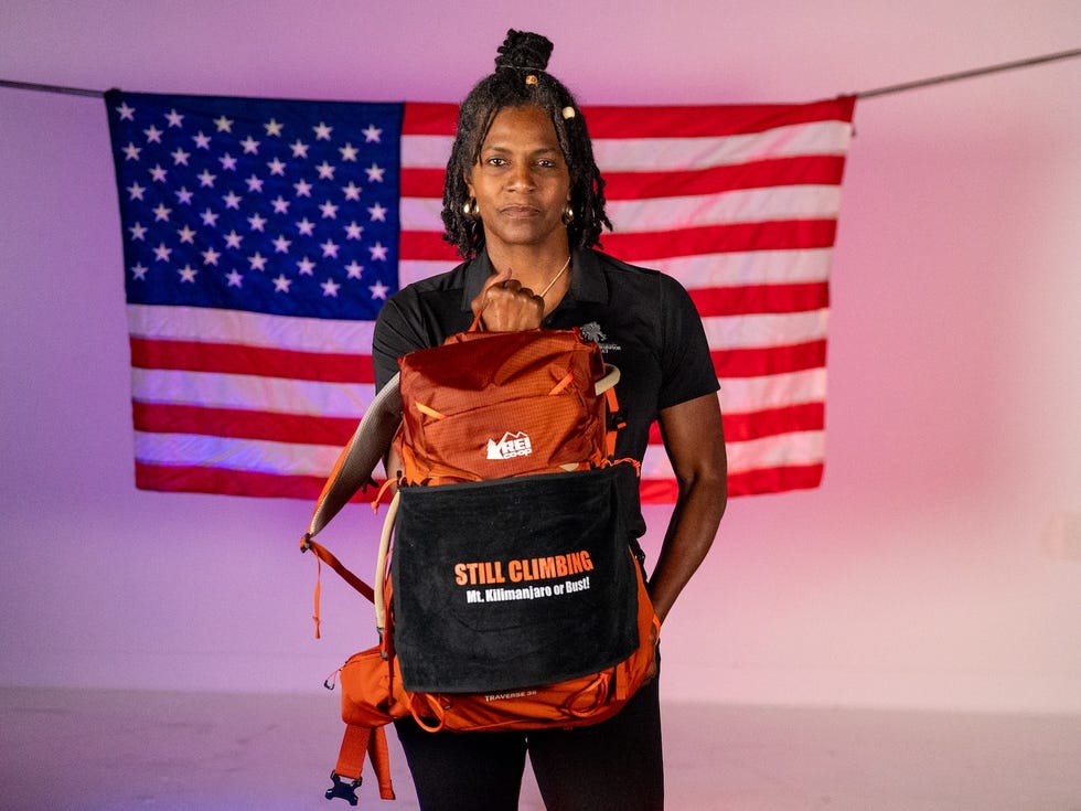 Person standing with a backpack featuring "STILL CLIMBING" text, against a backdrop of American flags. Person standing with a backpack featuring "STILL CLIMBING" text, against a backdrop of American flags.