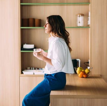 a young woman enjoys a peaceful moment with coffee in a contemporary kitchen setting featuring wooden cabinetry, green shelving accents, and natural lighting. perfect depiction of relaxation and modern interiors.