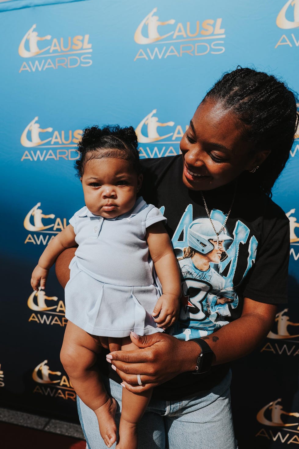 A smiling adult holding a baby in front of a blue backdrop with "CAUSL AWARDS" written on it. A smiling adult holding a baby in front of a blue backdrop with "CAUSL AWARDS" written on it.