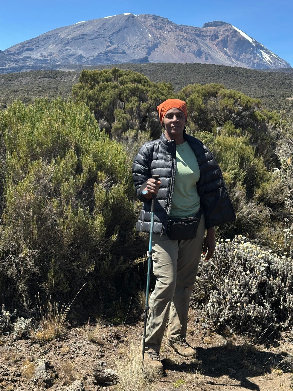 Hiker standing in front of a mountain, surrounded by shrubs, wearing a jacket and headscarf, holding a walking stick. Hiker standing in front of a mountain, surrounded by shrubs, wearing a jacket and headscarf, holding a walking stick.