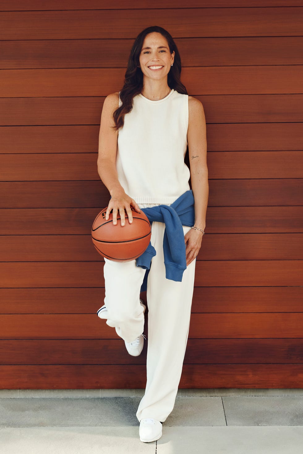 Person smiling, holding a basketball, leaning against a wooden wall.
