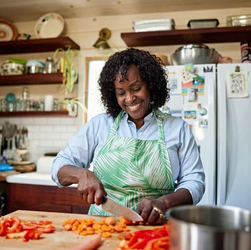 Smiling Black woman chopping vegetables in cozy kitchen waist up view of home cook in apron standing at cutting board and preparing red bell peppers and carrots for midday meal.
