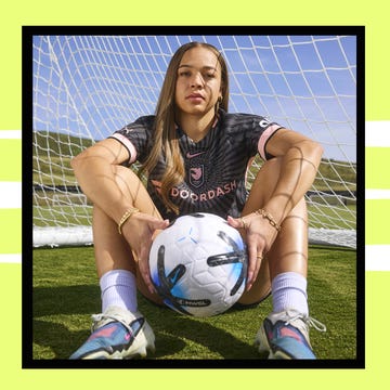 A soccer player sitting with a ball in front of a goal, wearing a team jersey and focused expression. A soccer player sitting with a ball in front of a goal, wearing a team jersey and focused expression.