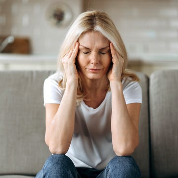 middle aged blonde woman sits on couch at living room holding her head with her hands, feels unhappy because of headache, personal troubles, illness or bad news, she need psychological or medical support