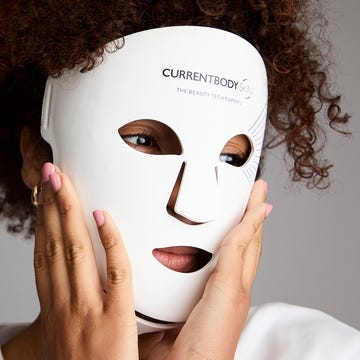 Person wearing a white LED facial mask with logo, touching their face, against a neutral gray background.