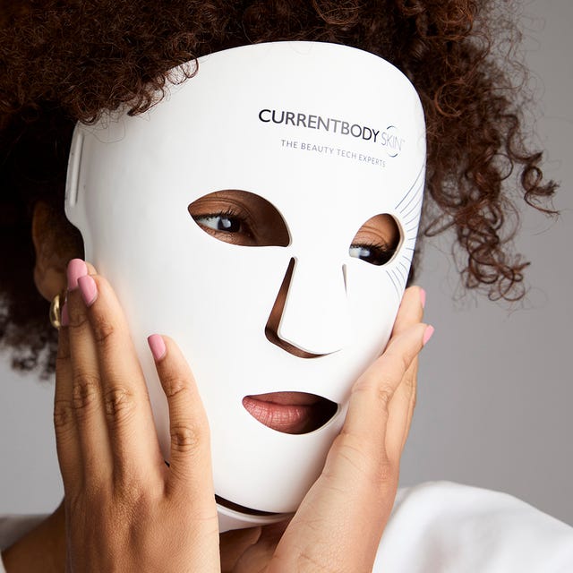 Person wearing a white LED facial mask with logo, touching their face, against a neutral gray background.