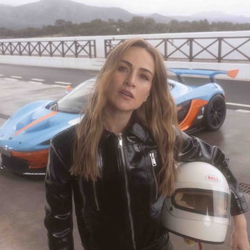 person in black leather jacket holding helmet by a blue and orange sports car on a race track.