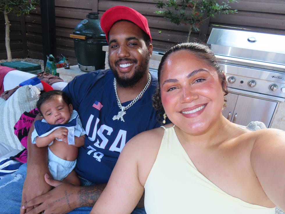 A smiling family together, with a man holding a baby, while a woman poses beside them in a casual outdoor setting. A smiling family together, with a man holding a baby, while a woman poses beside them in a casual outdoor setting.