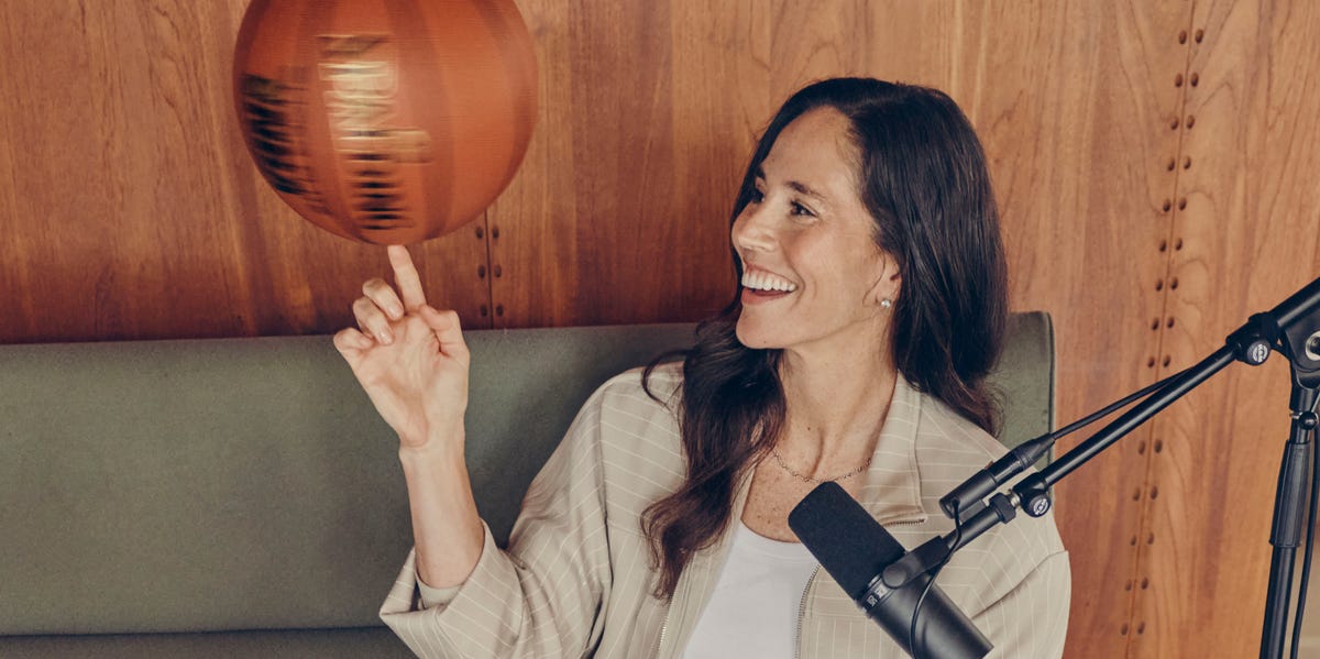Woman sitting on a couch, smiling, and spinning a basketball on her finger near a microphone.