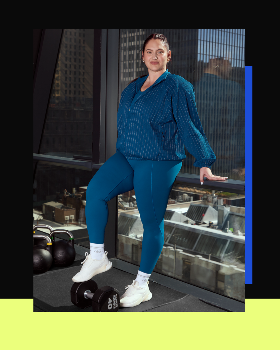 Woman in blue activewear in a gym next to dumbbells and kettlebells with a city view.