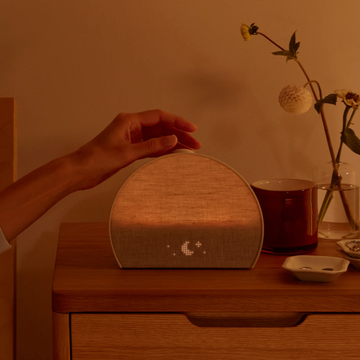 A hand adjusts a fabric-covered clock on a wooden nightstand with flowers and a mug nearby.