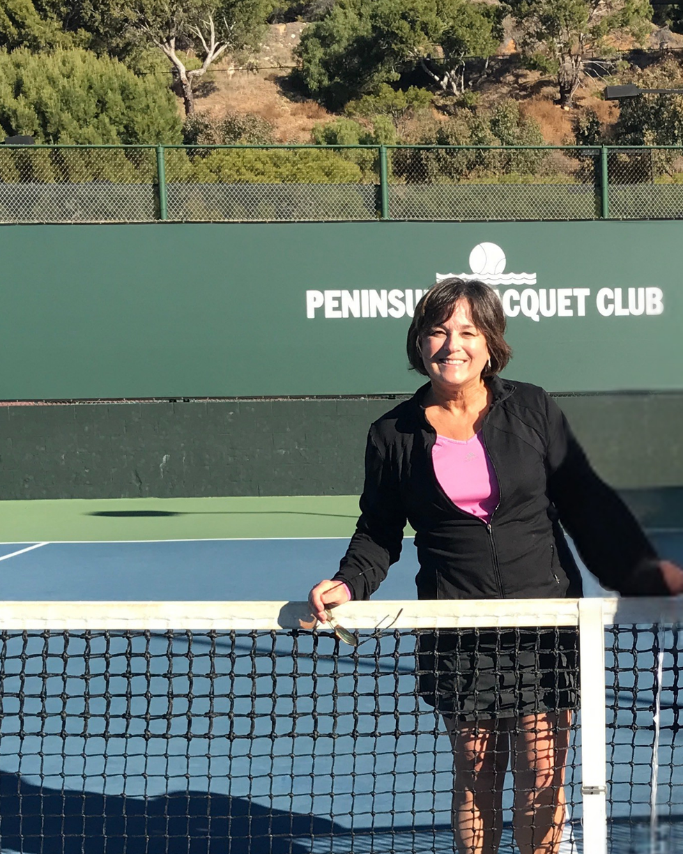 Smiling woman in casual tennis attire poses at the net of a tennis court with a club sign in the background.