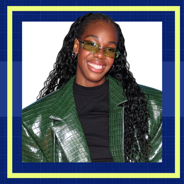 Smiling person with long, curly hair wearing a green textured jacket and sunglasses against a blue grid background.