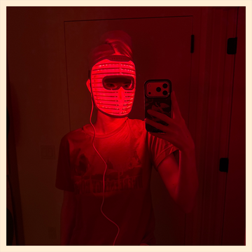 A light therapy mask in use, showing the user wearing it with red light illuminating the face in a bathroom setting.